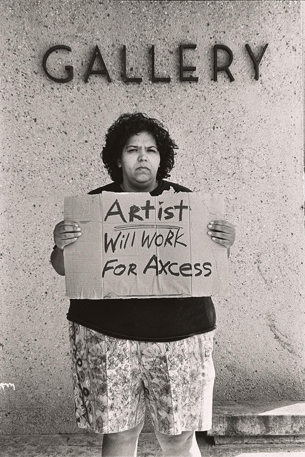 A grayscale photograph of a figure standing against a wall with the word “Gallery” above her head. Facing the camera, she holds a cardboard sign that reads “Artist will work for axcess,” spelled A-X-C-E-S-S.