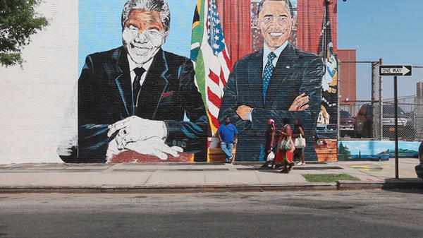 Painted wall mural of Barack Obama and Nelson Mandela along city street with several people walking in front.
